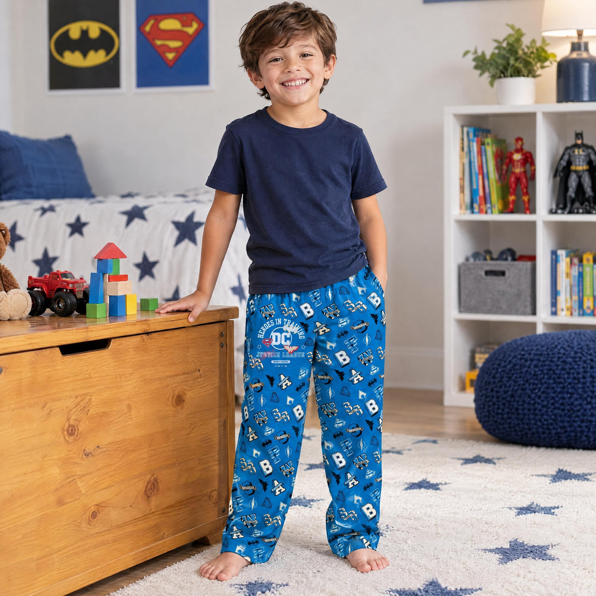 A child standing in a playroom wearing vibrant blue Justice League pajama pants with a comfortable, stretchy fit.