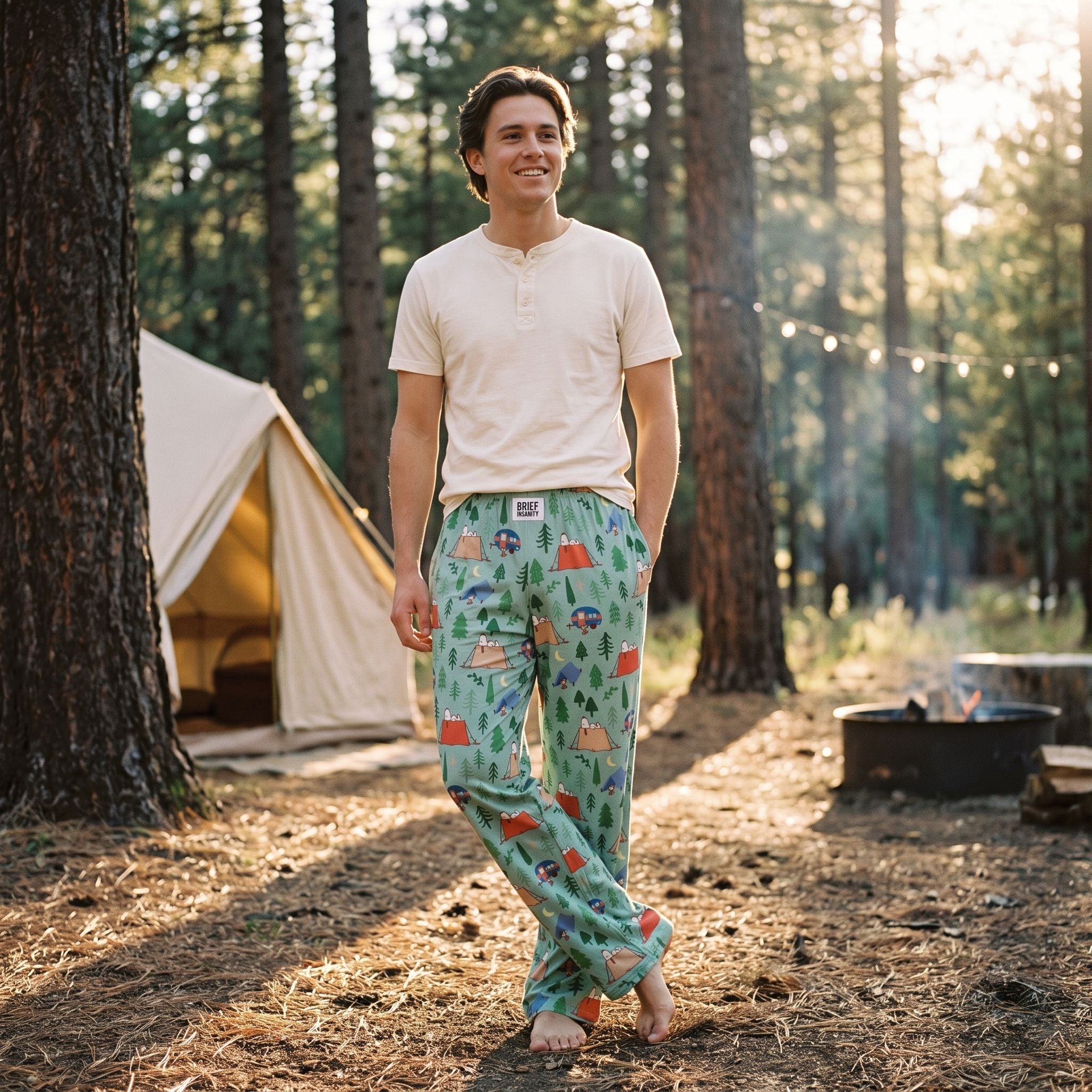 Young adult model relaxing outdoors in Snoopy camping-pattern lounge pants near a tent and pine trees.