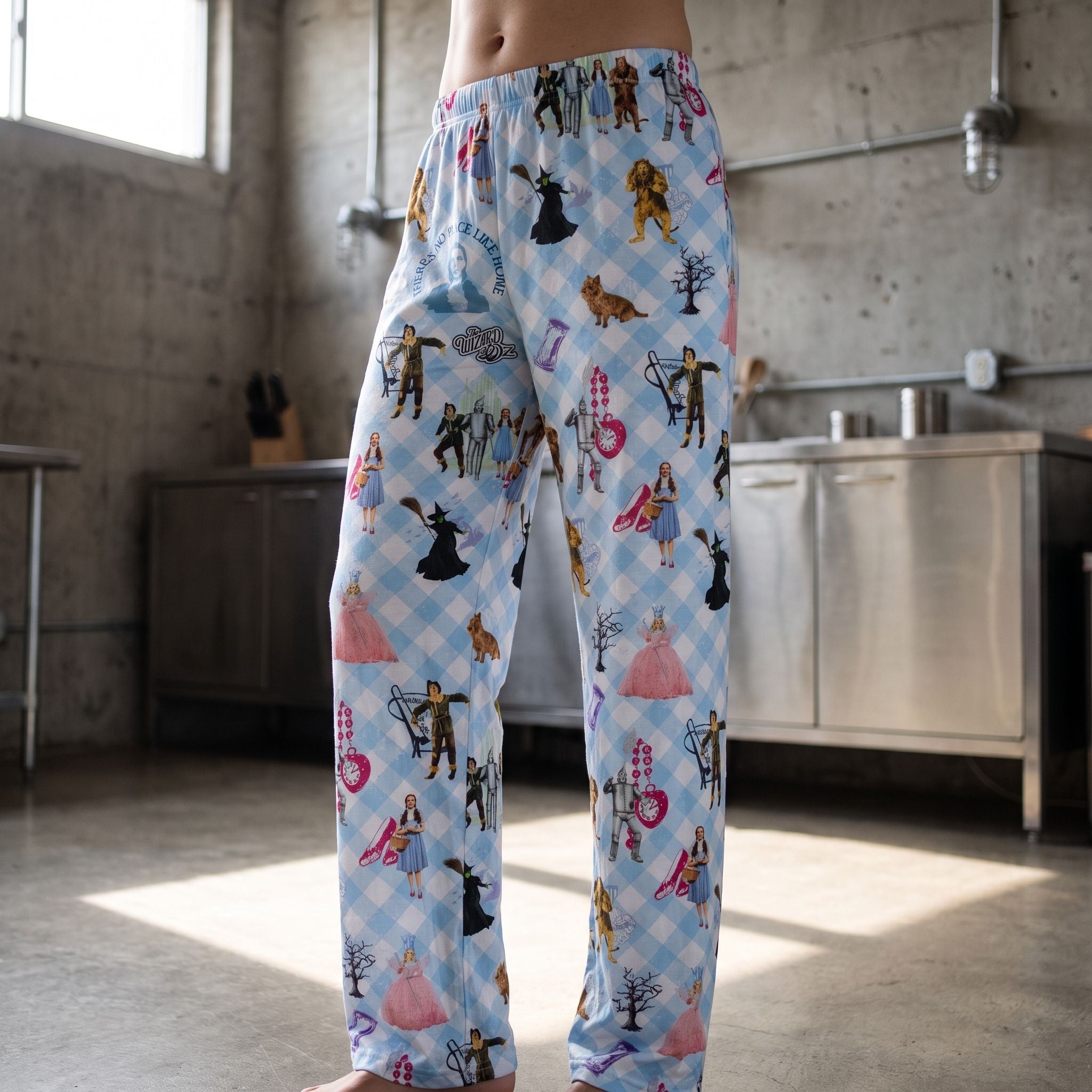 Model wearing Wizard of Oz lounge pants featuring iconic characters and gingham pattern in an industrial style kitchen.