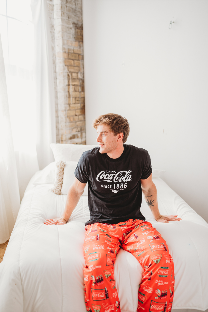 Image of a male model sitting on a white bed looking off into the distance wearing the Coca-Cola Retro Pattern pajama lounge pants