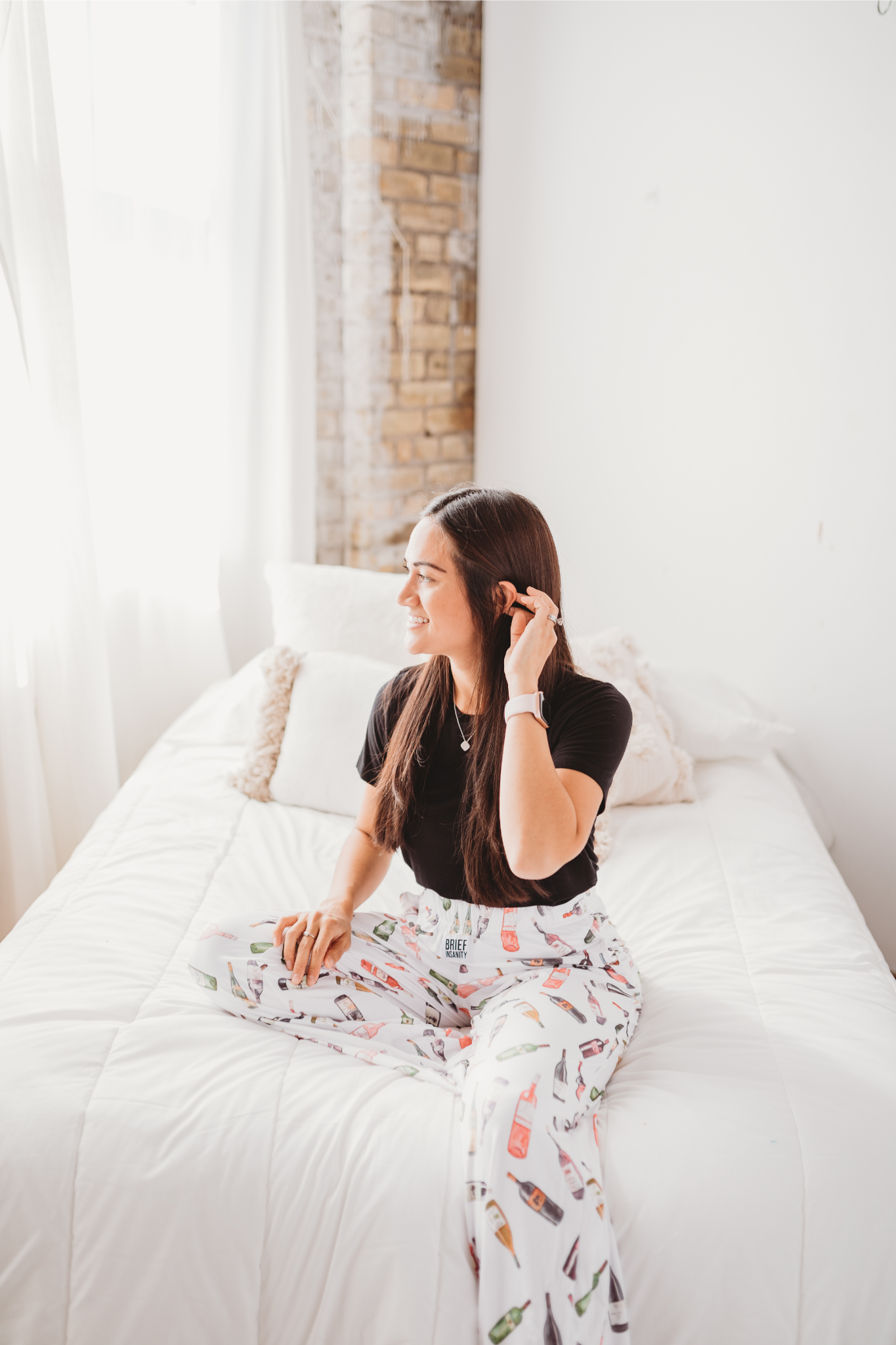 Female model sitting on white bed looking off to the side and smiling wearing Just Screw It Wine pajama lounge pants