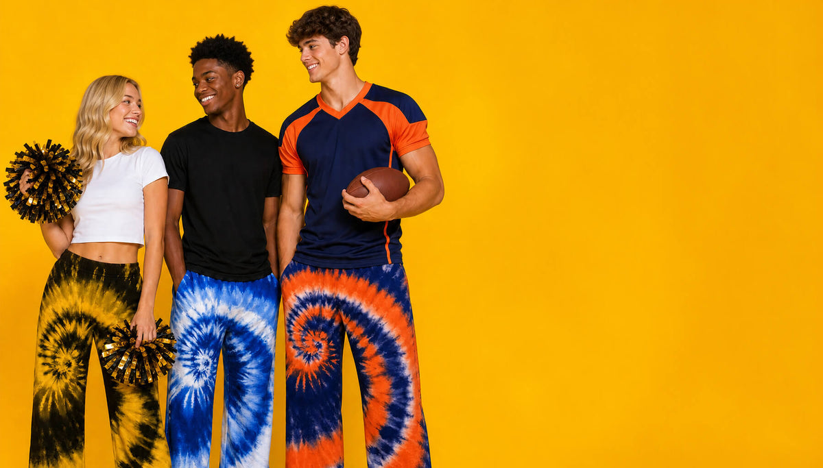Three models wearing matching tie-dye athletic sets with spiral patterns in blue, orange, black, and white, standing against a solid yellow background. One model holds a football, and two hold cheerleader pom-poms.