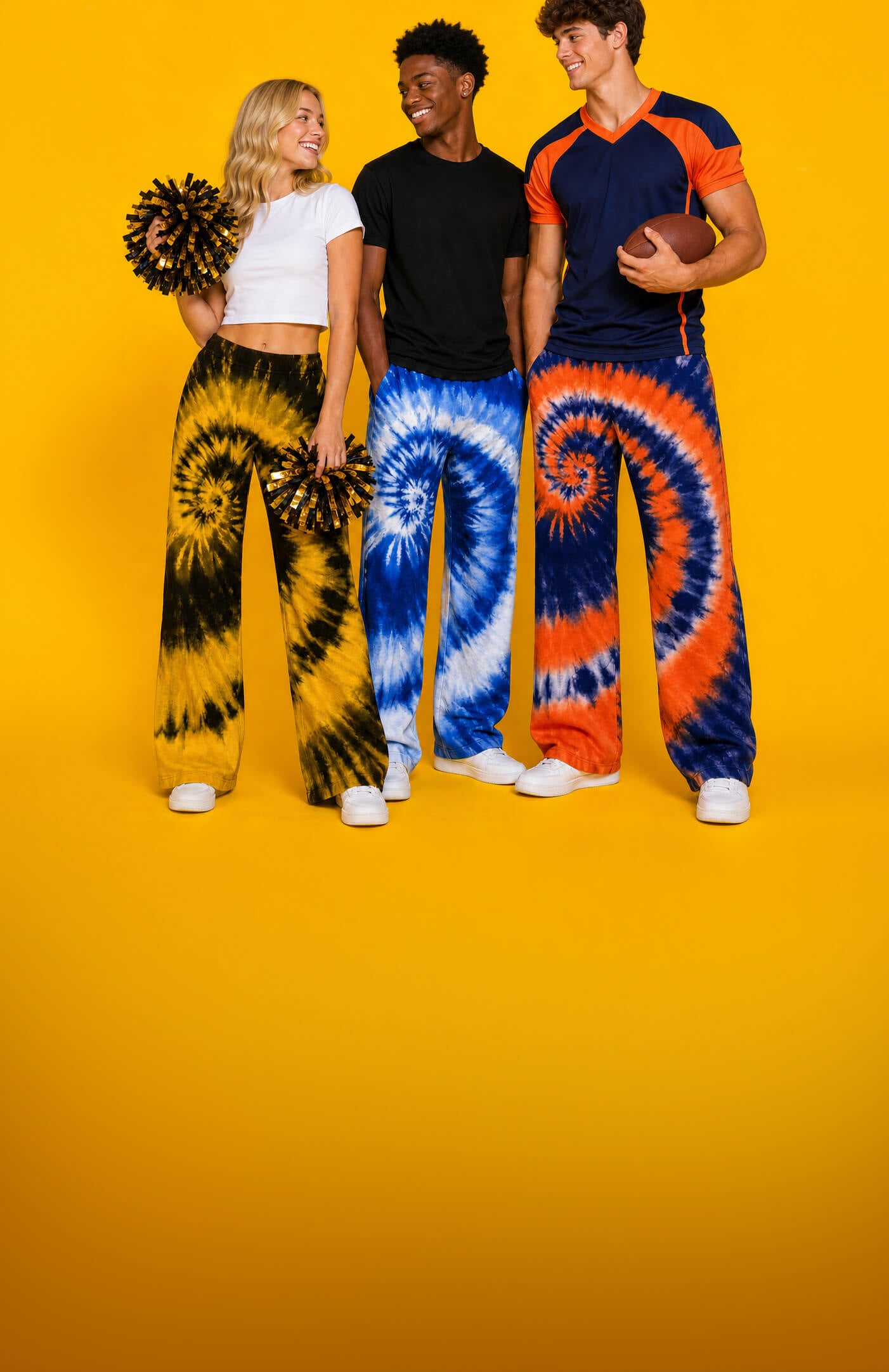 Three models standing against a solid yellow studio background, wearing matching tie-dye wide-leg sweatpants in black/yellow, blue/white, and orange/blue spiral patterns, with cheer pom-poms and a football.