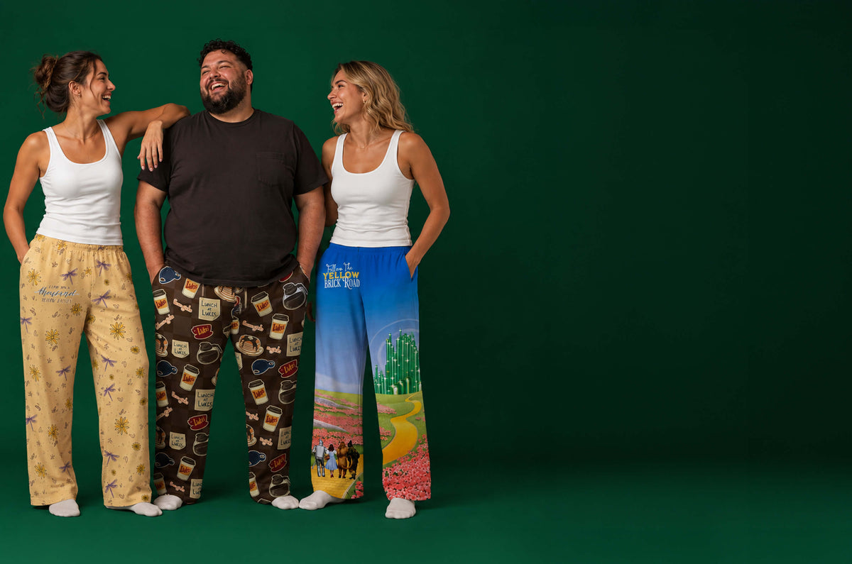 Three models wearing colorful novelty pajama pants for Gilmore Girls and WIzard of Oz stand against a solid dark green studio background. The pants feature various designs: one with a coffee cup pattern, one with a detailed landscape print, and one with a floral print.
