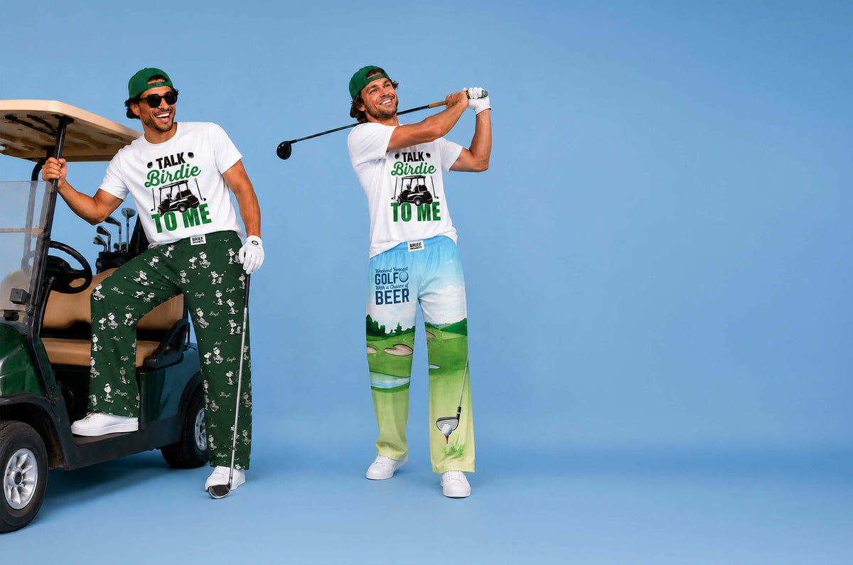 Two men modeling matching white graphic t-shirts and green patterned golf pants against a solid blue studio background. One man is sitting in a golf cart, and the other is mid-swing with a golf club.