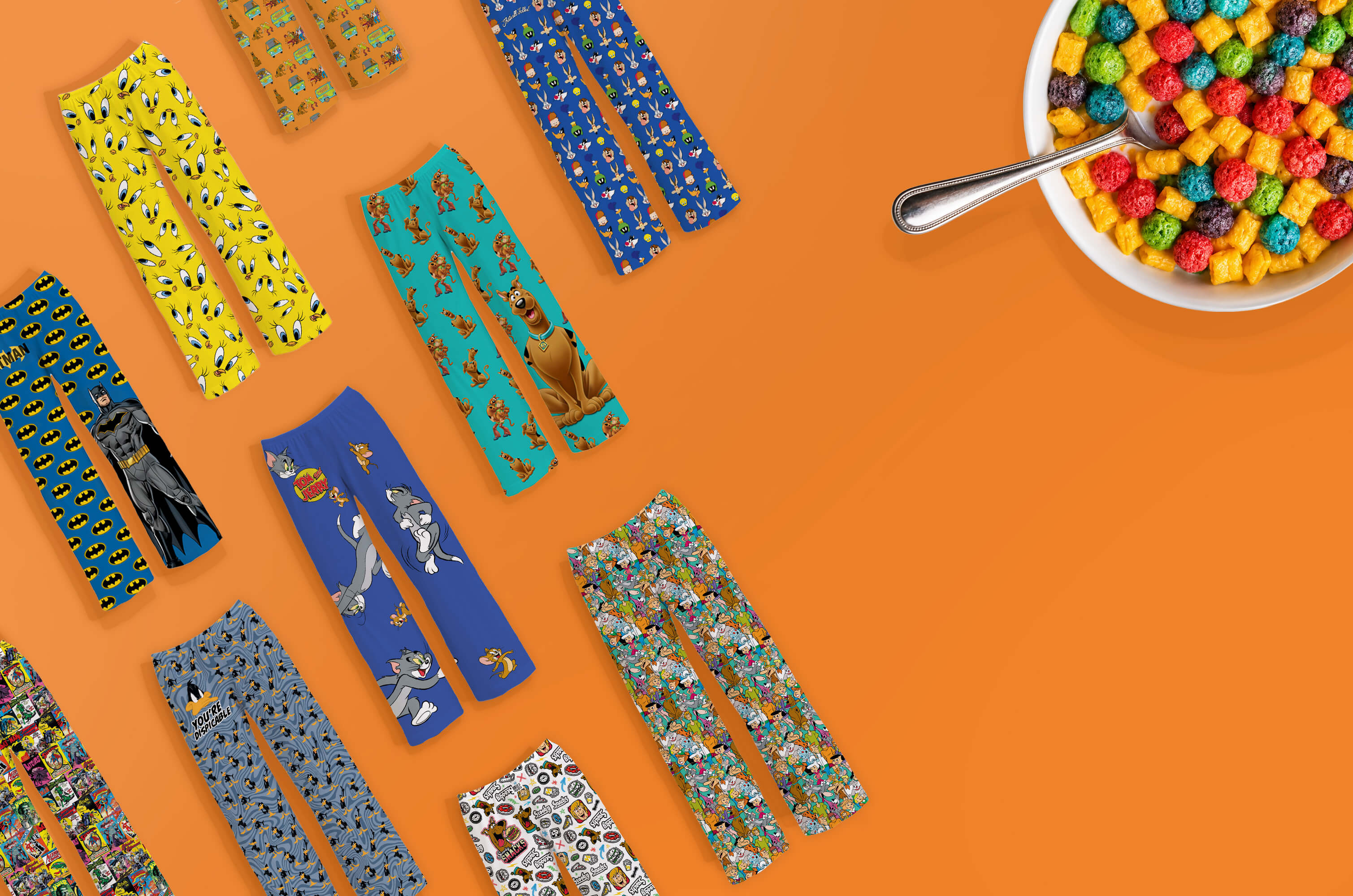 A styled flat-lay of multiple pairs of colorful Saturday Morning Cartoon patterned pants arranged on a bright orange background, with a bowl of multicolored cereal and a spoon in the top-right corner.