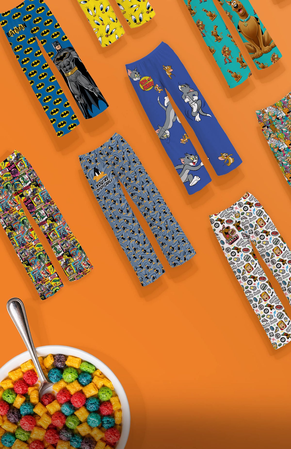 A styled flat-lay of multiple pairs of colorful Saturday Morning Cartoon patterned pants arranged on a bright orange background, with a bowl of multicolored cereal and a spoon in the top-right corner.A styled flat-lay of multiple pairs of colorful Saturday Morning Cartoon patterned pants arranged on a bright orange background, with a bowl of multicolored cereal and a spoon in the top-right corner.