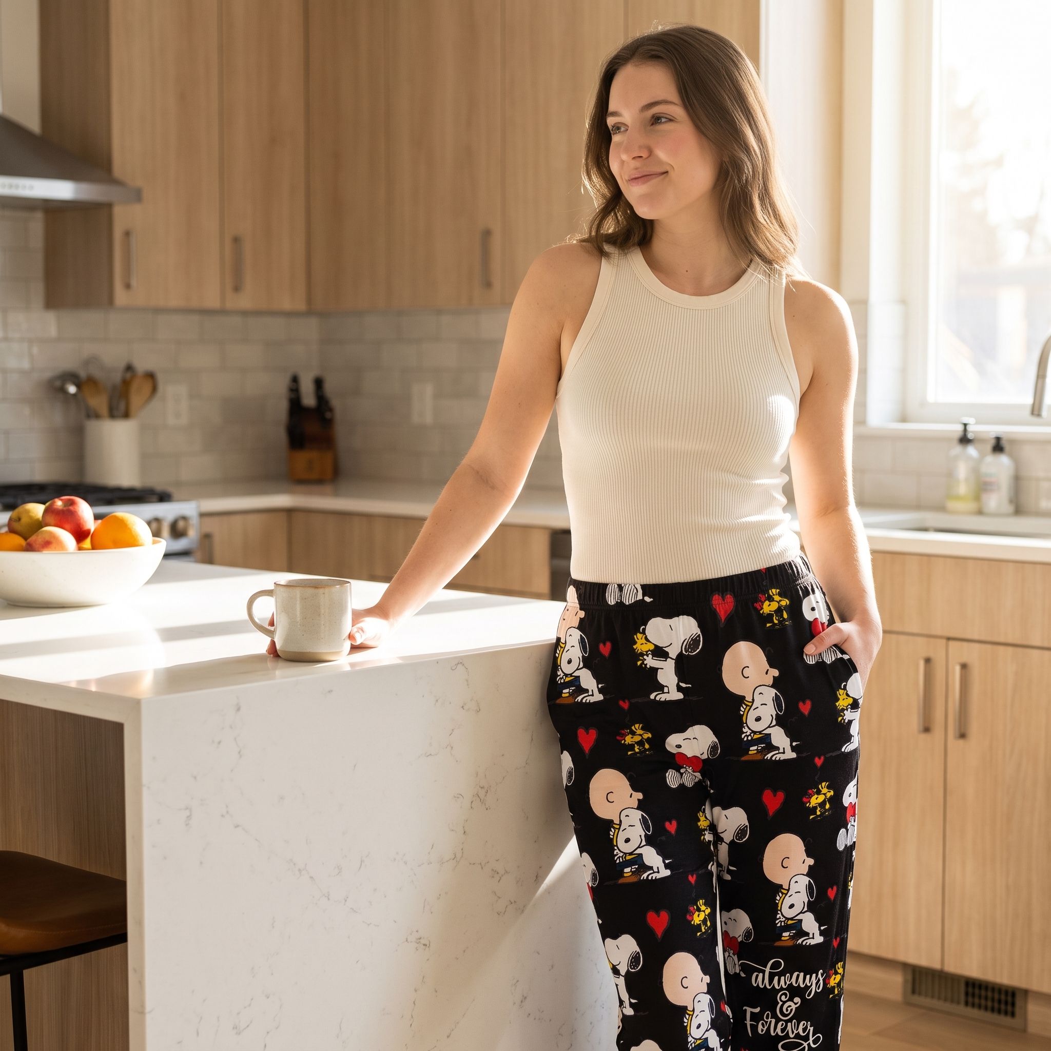Casual woman wearing Snoopy and hearts lounge pants from the Always & Forever collection, relaxed at home or in a kitchen with morning sunlight.