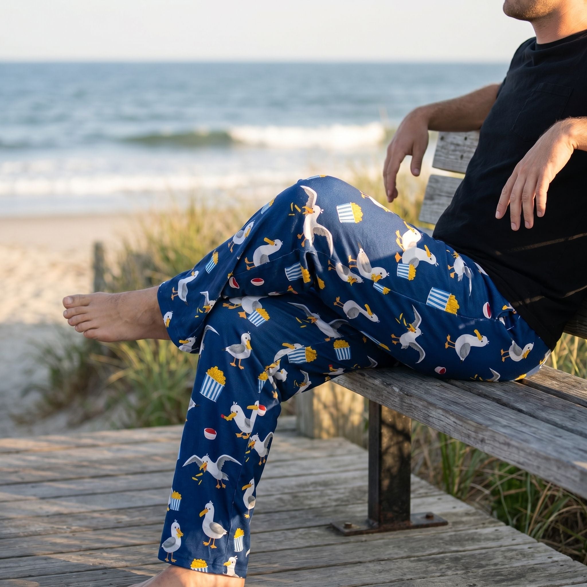 Relaxed model wearing Seagull French Fry Lounge Pants with playful seagull print, sitting on weathered wooden bench near coast, natural sunlight.