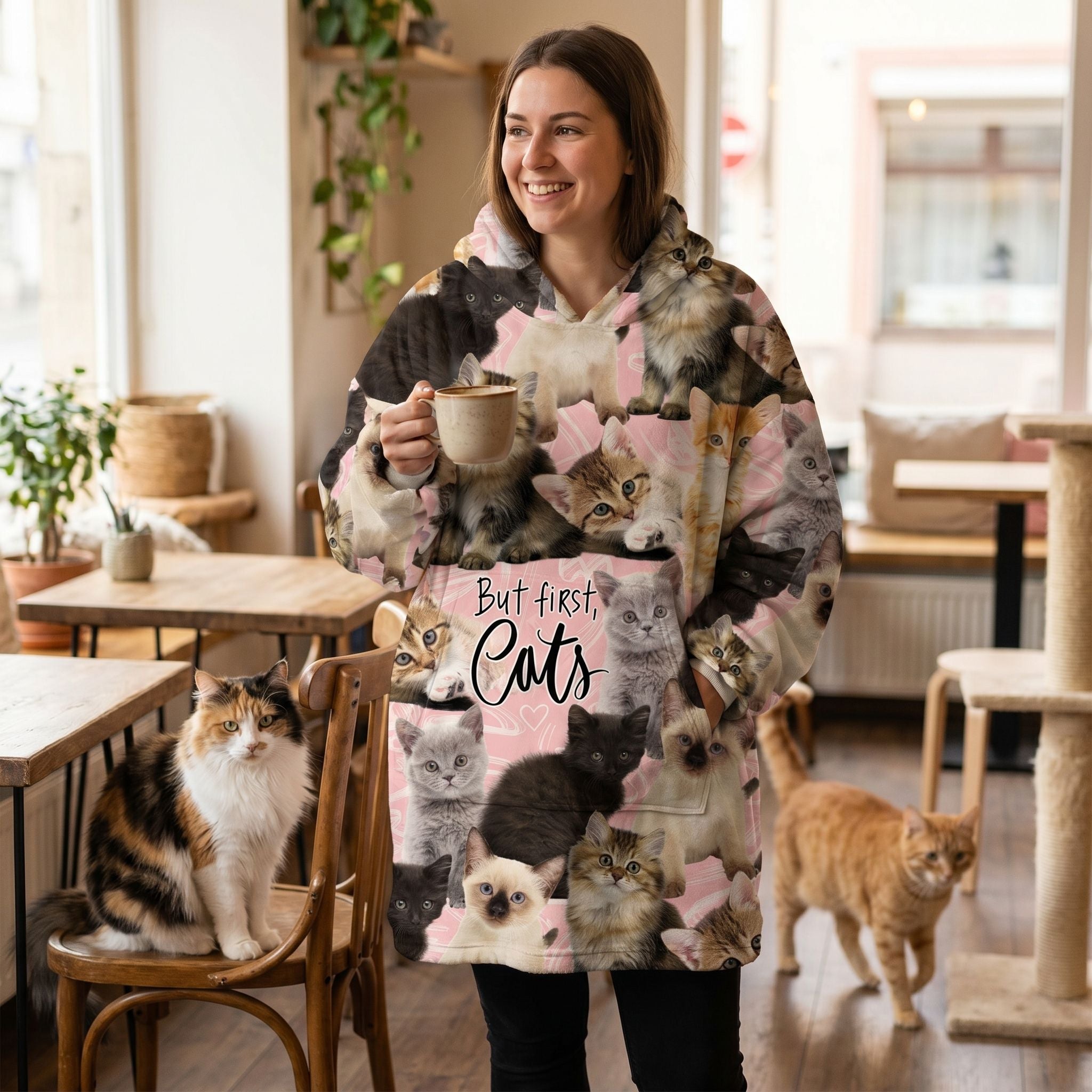 Woman smiling in a cat café wearing an oversized blanket hoodie with kitten designs, surrounded by friendly cats and warm lighting.