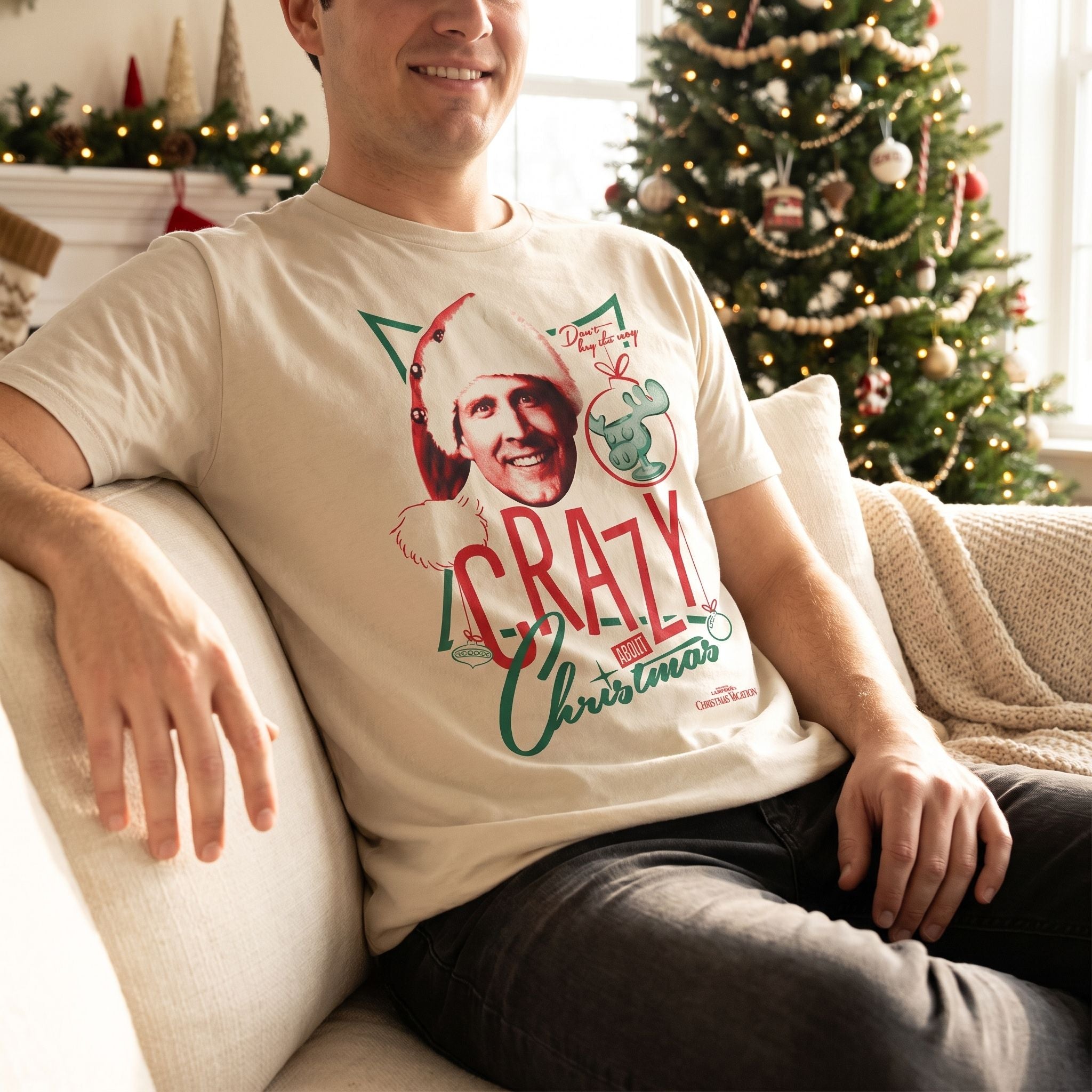 Model wearing Crazy Christmas Lampoon's Christmas Vacation Shirt sitting on a plush sofa with Christmas decorations and soft lighting