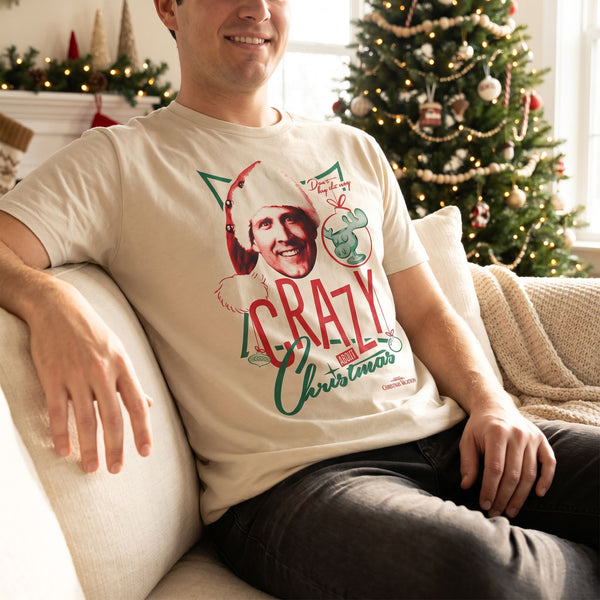 Model wearing Crazy Christmas Lampoon's Christmas Vacation Shirt sitting on a plush sofa with Christmas decorations and soft lighting
