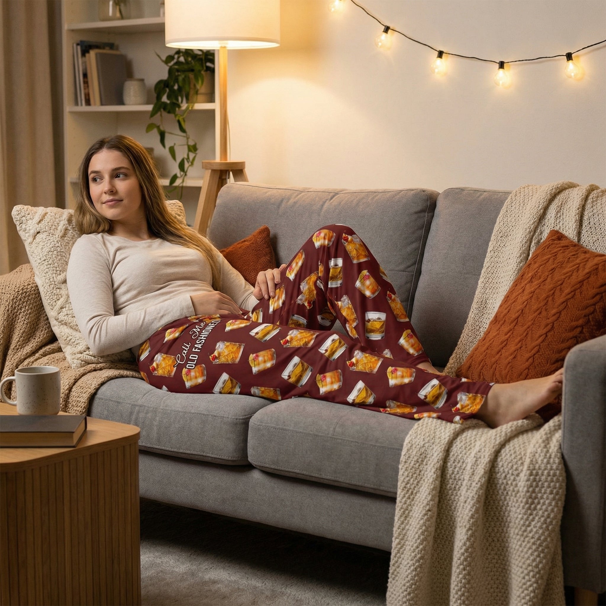 Relaxed woman lounging on sofa in cozy living room, wearing Old Fashioned Lounge Pants with vivid cocktail print, warm ambient light.