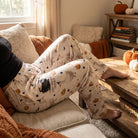 Whimsical Brewin' Trouble & Gooses lounge pants worn by a model in a cozy living room, showing pumpkin and witch hat pattern, elastic waistband, and pockets.