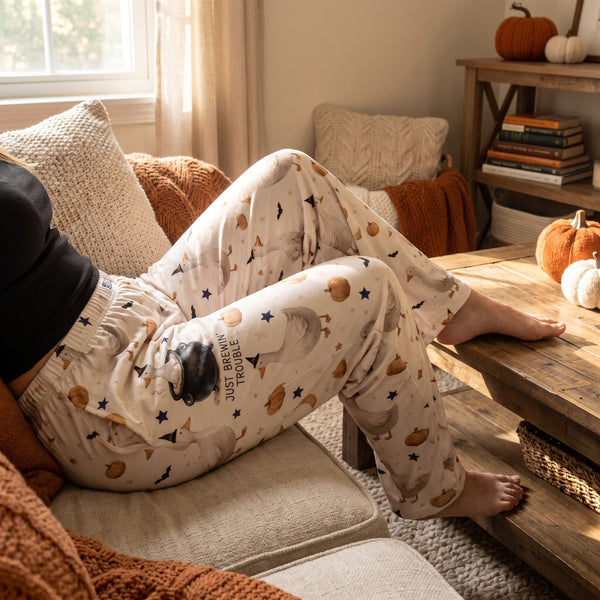 Whimsical Brewin' Trouble & Gooses lounge pants worn by a model in a cozy living room, showing pumpkin and witch hat pattern, elastic waistband, and pockets.