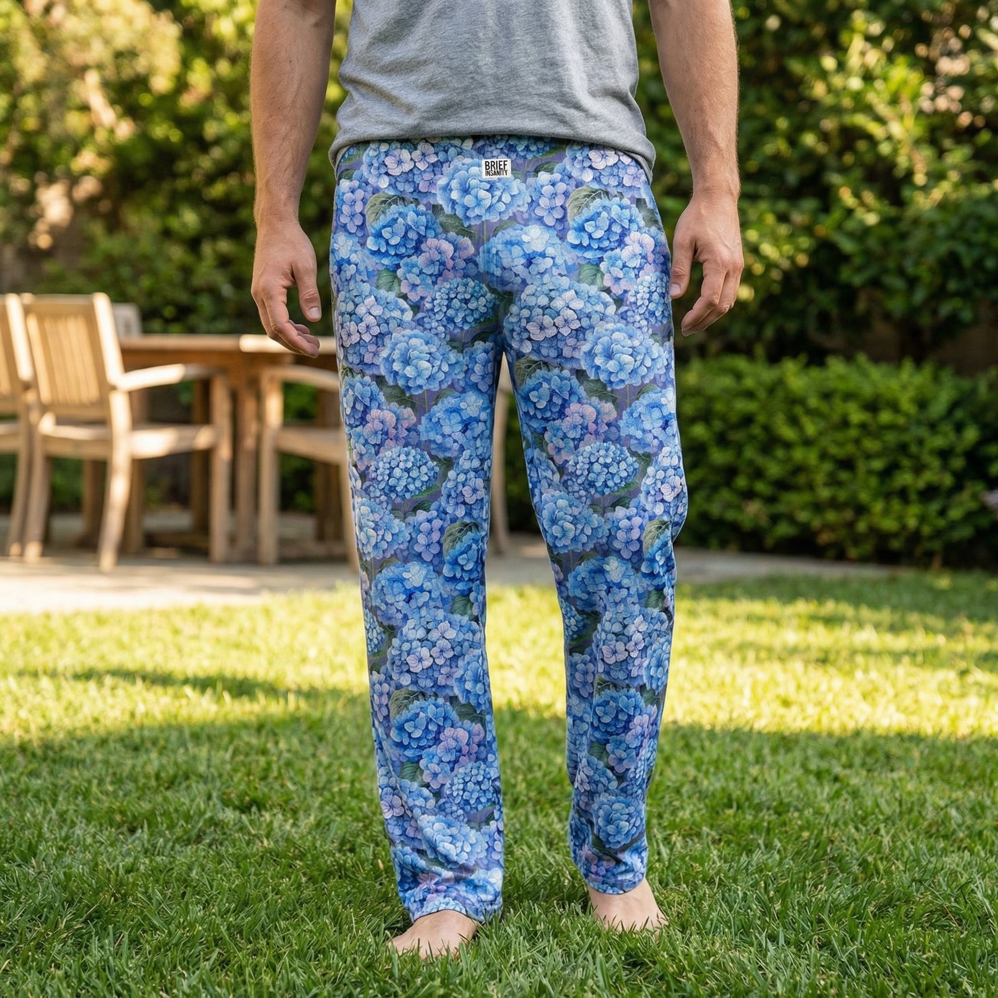 Female model standing outdoors in a sunny backyard wearing blue hydrangea lounge pants and a neutral t-shirt, relaxed and natural pose, pants clearly visible.