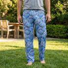 Female model standing outdoors in a sunny backyard wearing blue hydrangea lounge pants and a neutral t-shirt, relaxed and natural pose, pants clearly visible.