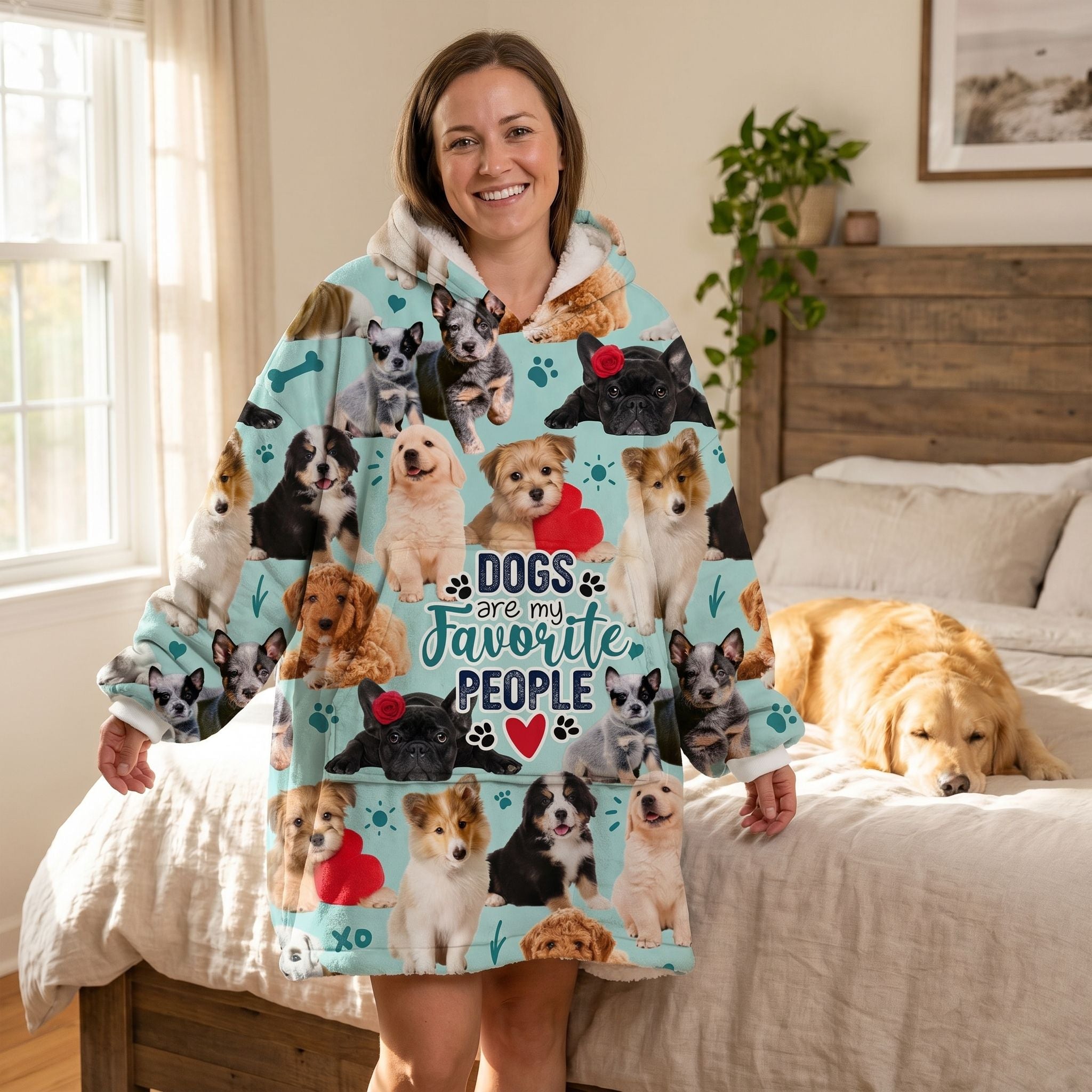 Lifestyle image of smiling woman in oversized dog-themed blanket hoodie standing in cozy bedroom with dog resting on bed.