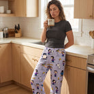 Adult female model wearing Snoopy Peanuts lounge pants, standing in bright modern kitchen, relaxed and holding coffee mug.