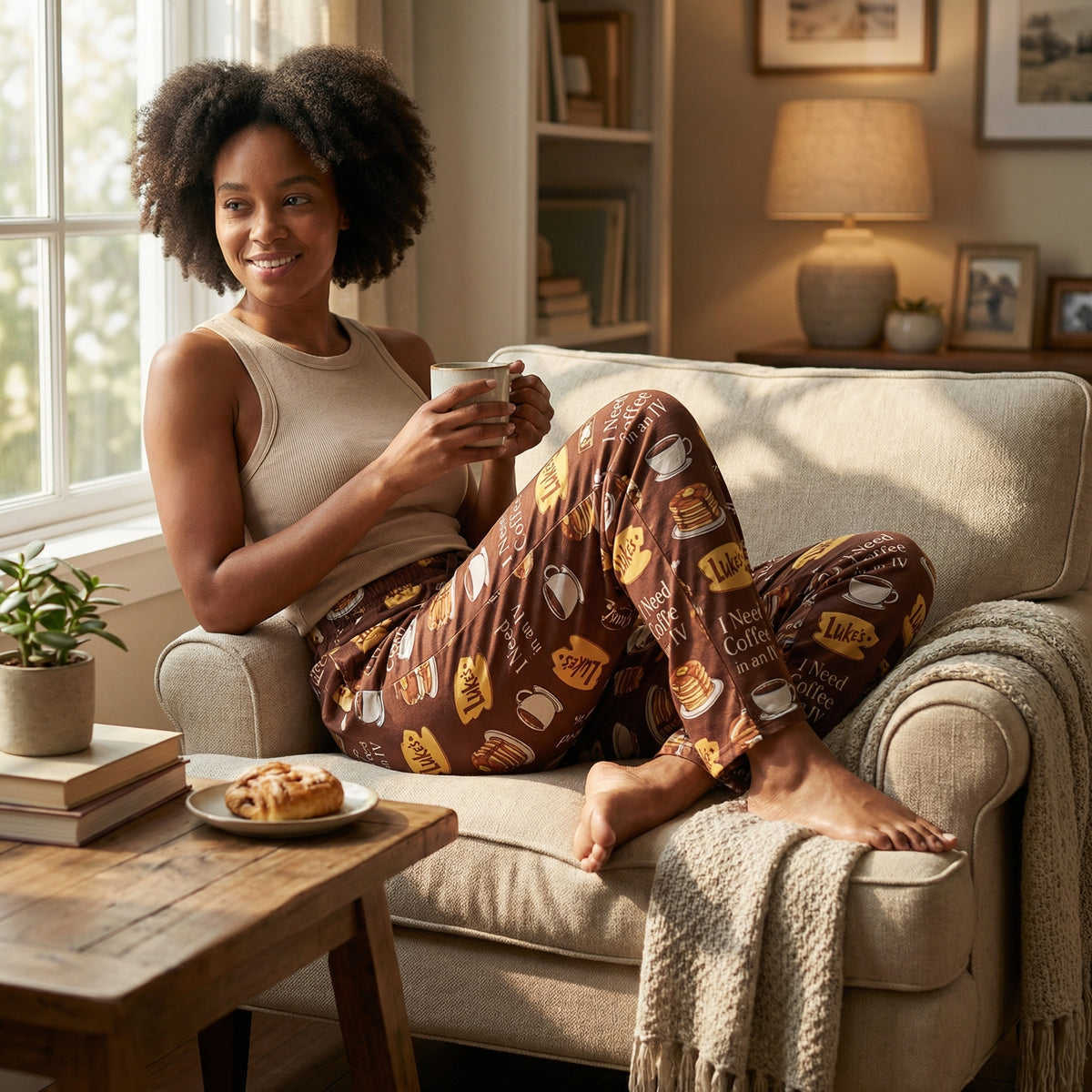 Female model relaxing in cozy living room wearing Luke's Coffee Diner Notes Gilmore Girls lounge pants with coffee mug and sunlight.
