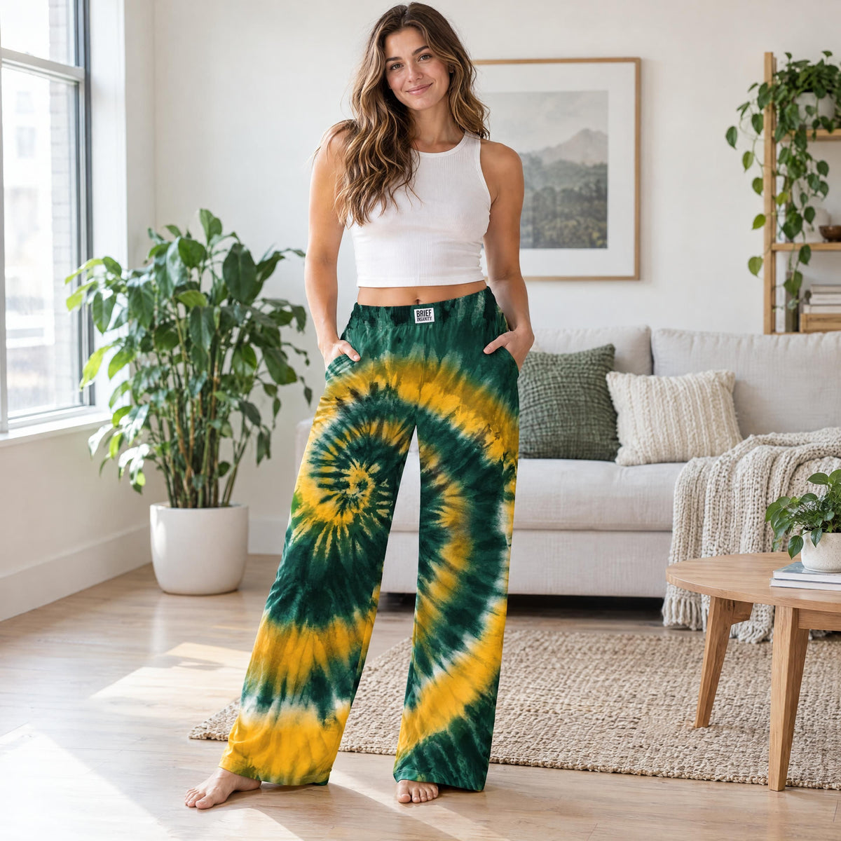 A person standing in a bright living room wearing wide leg lounge pants with a green and yellow spiral tie dye design.