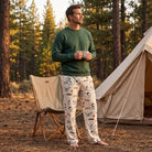 Male model standing at campsite in Snoopy camping badge lounge pants, holding mug near tent and pine trees during golden hour outdoor scene.