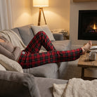 Model lounging on a modern sofa wearing red plaid pajama pants in a cozy living room scene.