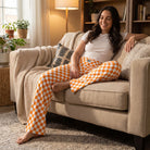 Full-body of a model relaxing in orange checker lounge pants seated on a plush sofa, showcasing their comfortable fit and bright colors.