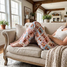 Person lounging on vintage farmhouse sofa wearing Highland Cow Quilt pajama pants with playful patchwork design.