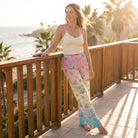 Young woman standing on sunlit balcony wearing pastel ombré Snoopy beach lounge pants with cream camisole, enjoying golden hour sunlight.
