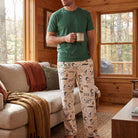 Adult male model wearing Snoopy camping badge lounge pants, relaxing in cozy cabin with natural wood details and large window scene.