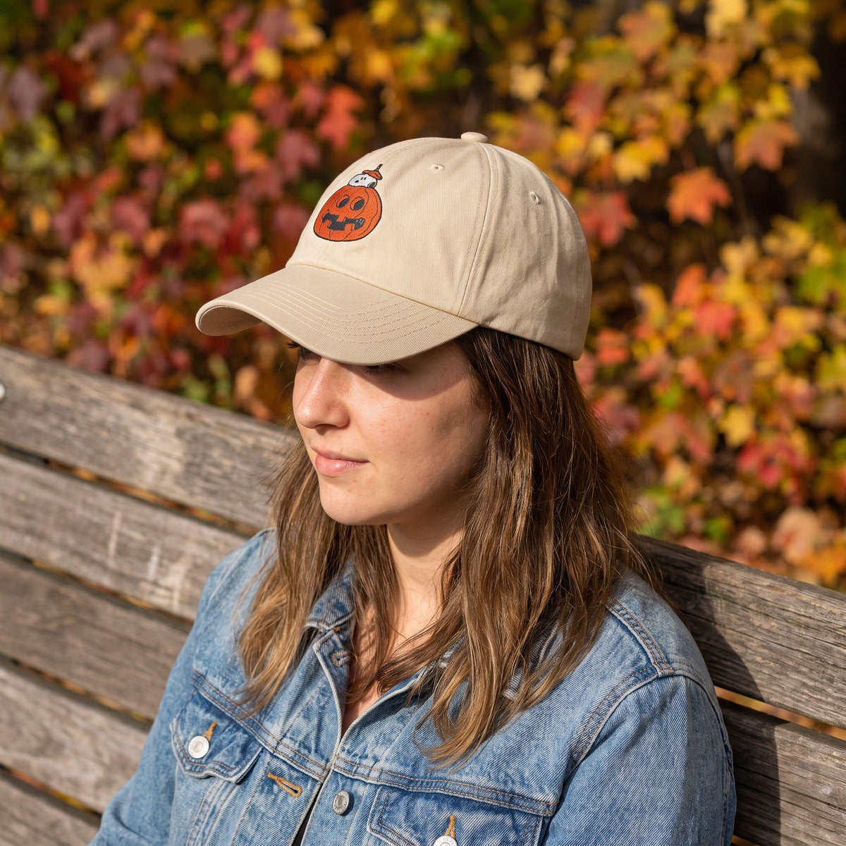 Model wearing Snoopy in a pumpkin embroidered cap on a park bench amid autumn leaves, highlighting the cap's fit and design.