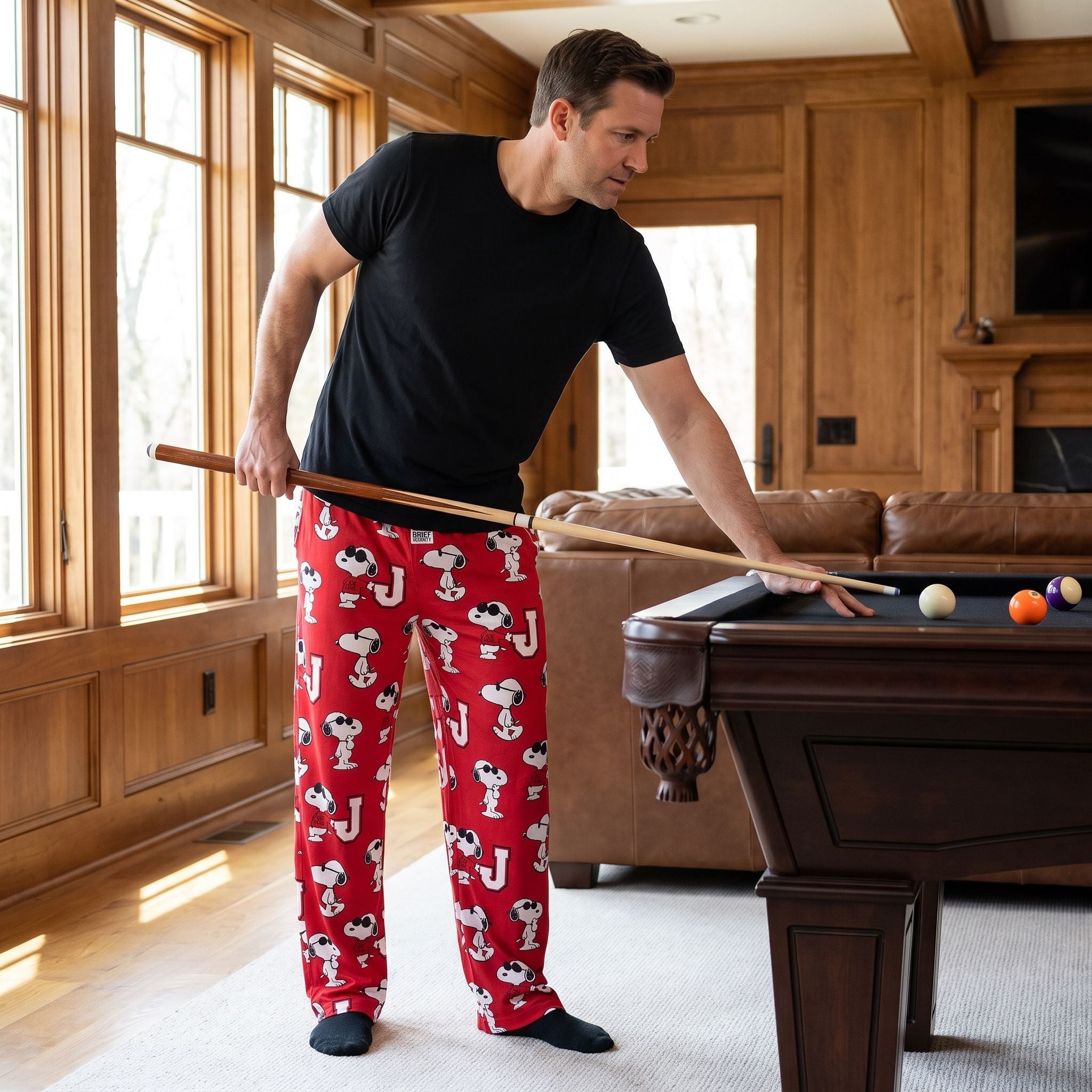 Adult male model in red Snoopy Joe Cool lounge pants standing beside a pool table in a cozy home recreation room.
