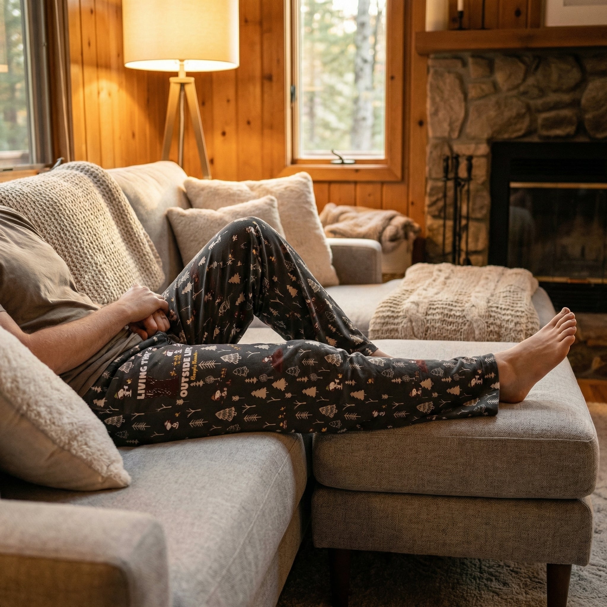 Model relaxing on a modern couch in a cozy cabin, showcasing Snoopy Outside Life lounge pants with woodland and campsite print.