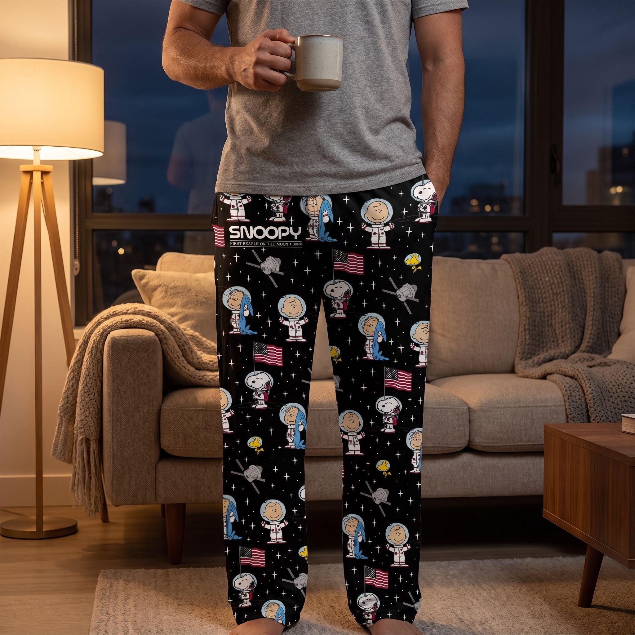 Adult male model wearing Snoopy Space Lounge Pants in a cozy living room, standing beside a sofa with a mug, night sky visible through windows.