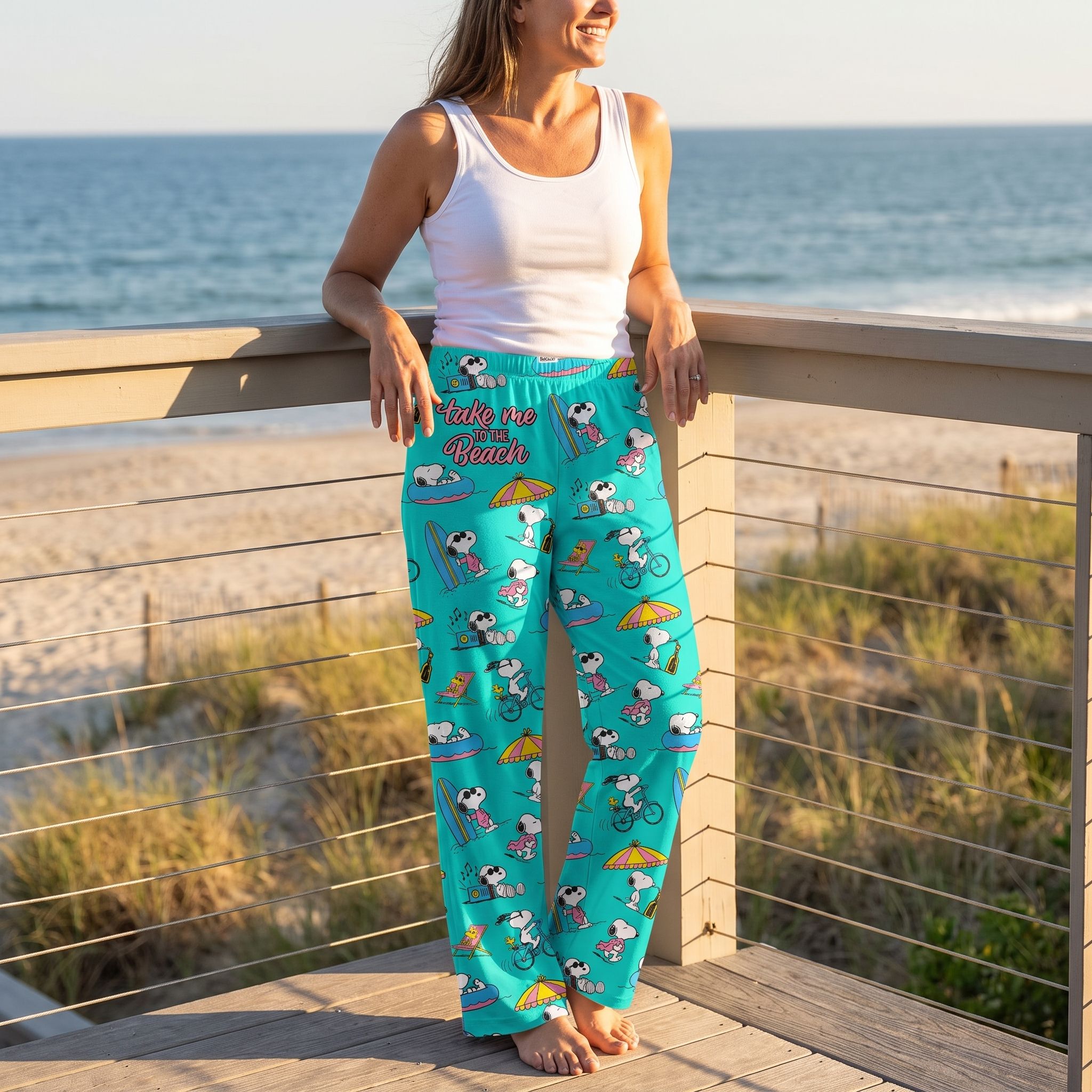 Model in Snoopy beach lounge pants on a sunny beach house balcony with ocean view, relaxed pose, showcasing product details.