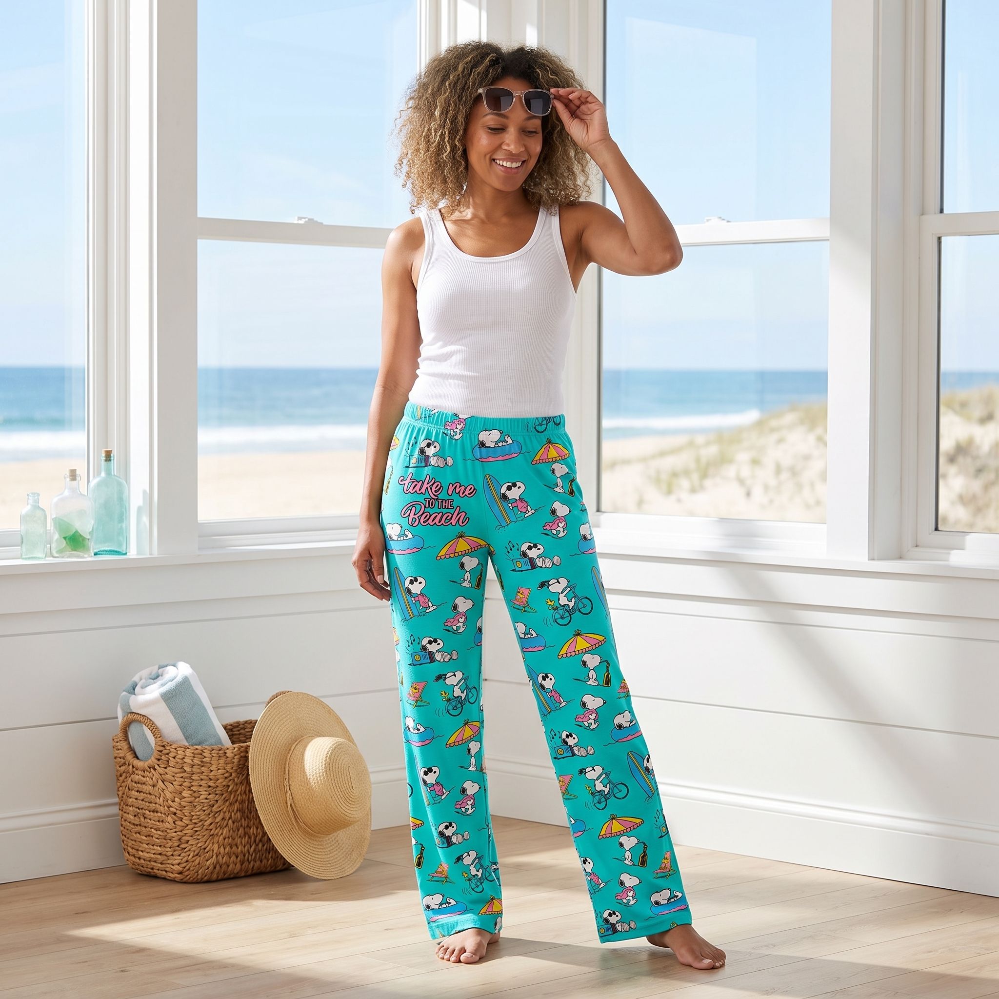 Adult female model wearing turquoise Snoopy beach lounge pants in a bright coastal beach house setting, natural sunlight, full-length view.