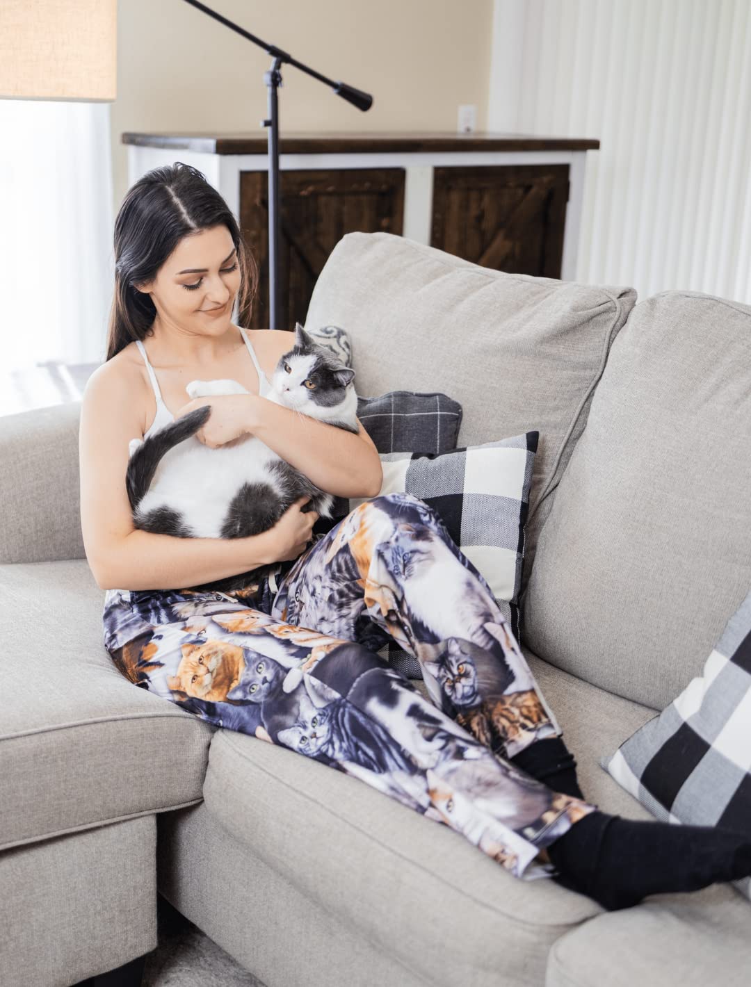 Image of female model sitting on gray couch and holding a big fluffy cat wearing All Over Cat pajama lounge pants (front view)