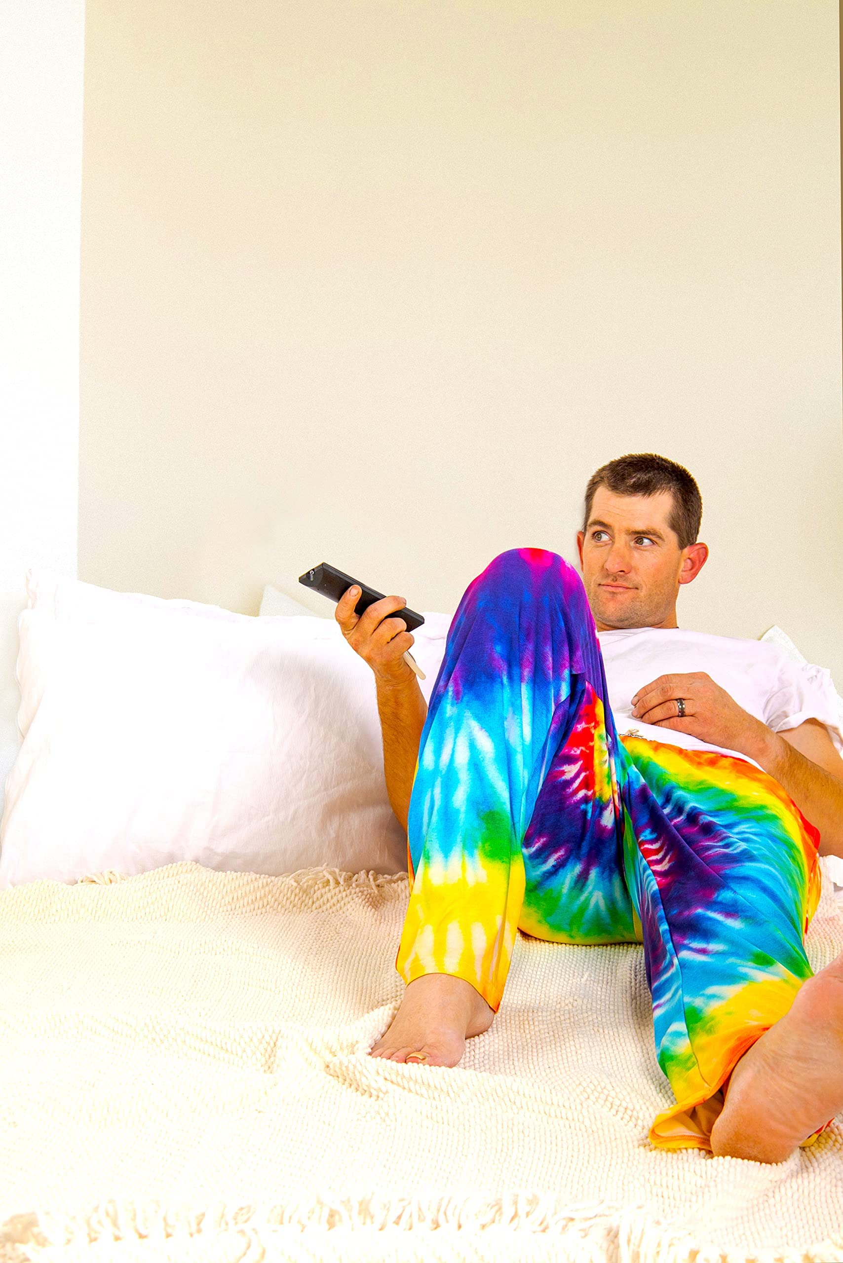 Image of male model laying down on bed with remote in hand wearing Rainbow Tie-Dye pajama lounge pants (front view)