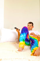 Image of male model laying down on bed with remote in hand wearing Rainbow Tie-Dye pajama lounge pants (front view)