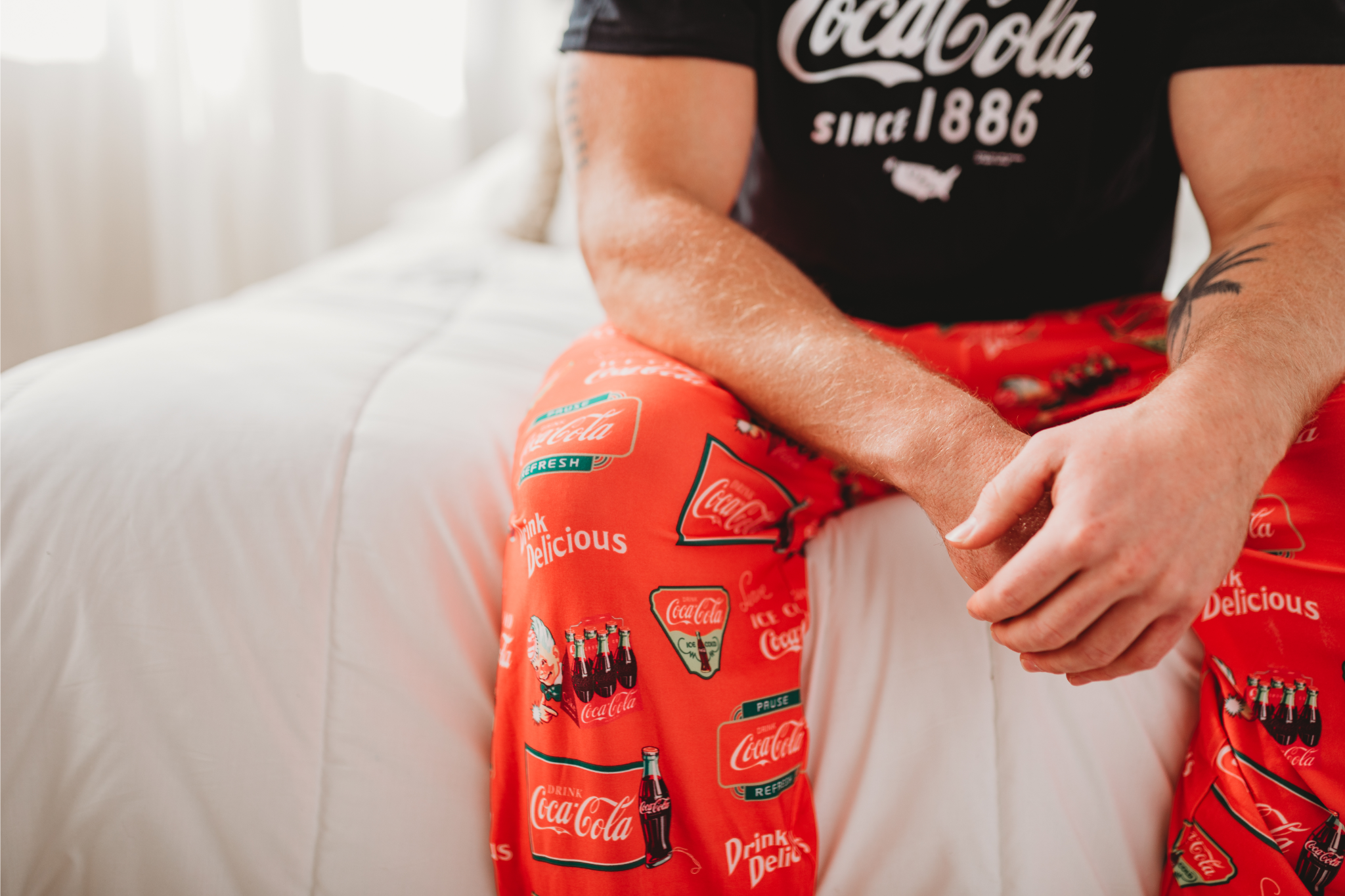 Close up image of a male model sitting on a bed wearing the Coca-Cola Retro Pattern pajama lounge pants