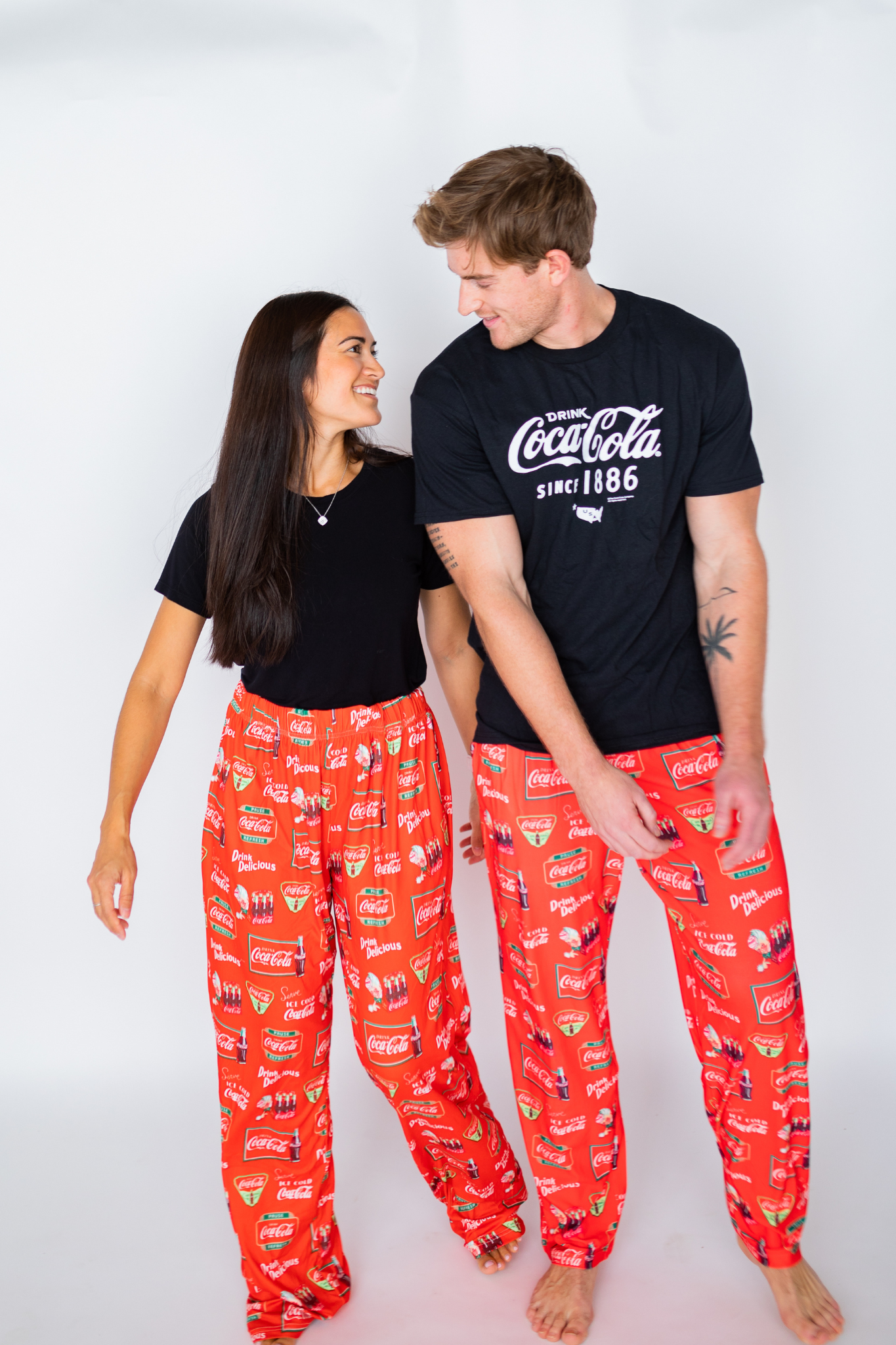 Image of female model and male model standing next to each other looking and smiling at each other both wearing the Coca-Cola Retro Pattern pajama lounge pants