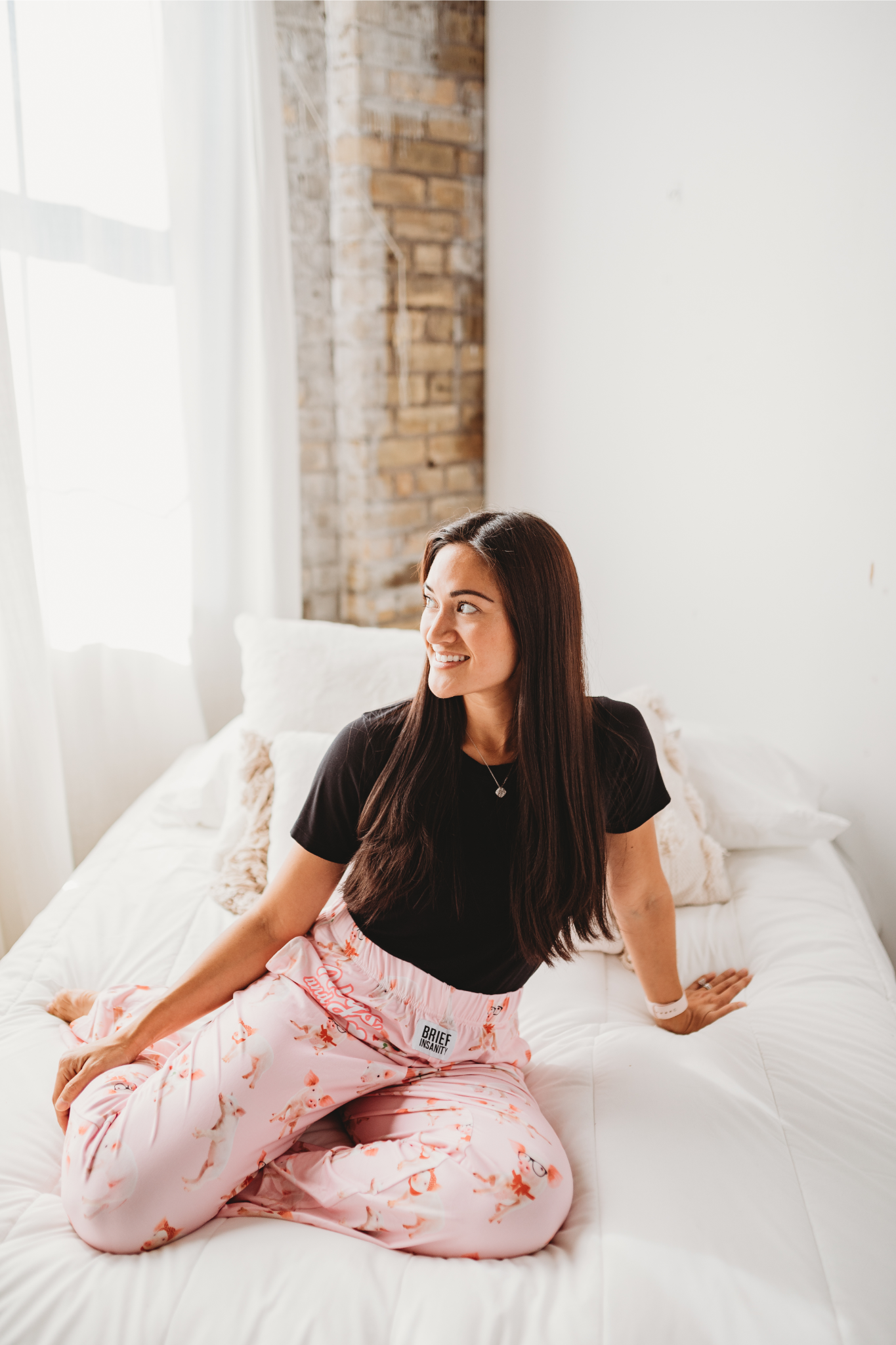 Female model sitting on white bed looking off into the distance smiling wearing Hogs And Kisses pajama lounge pants