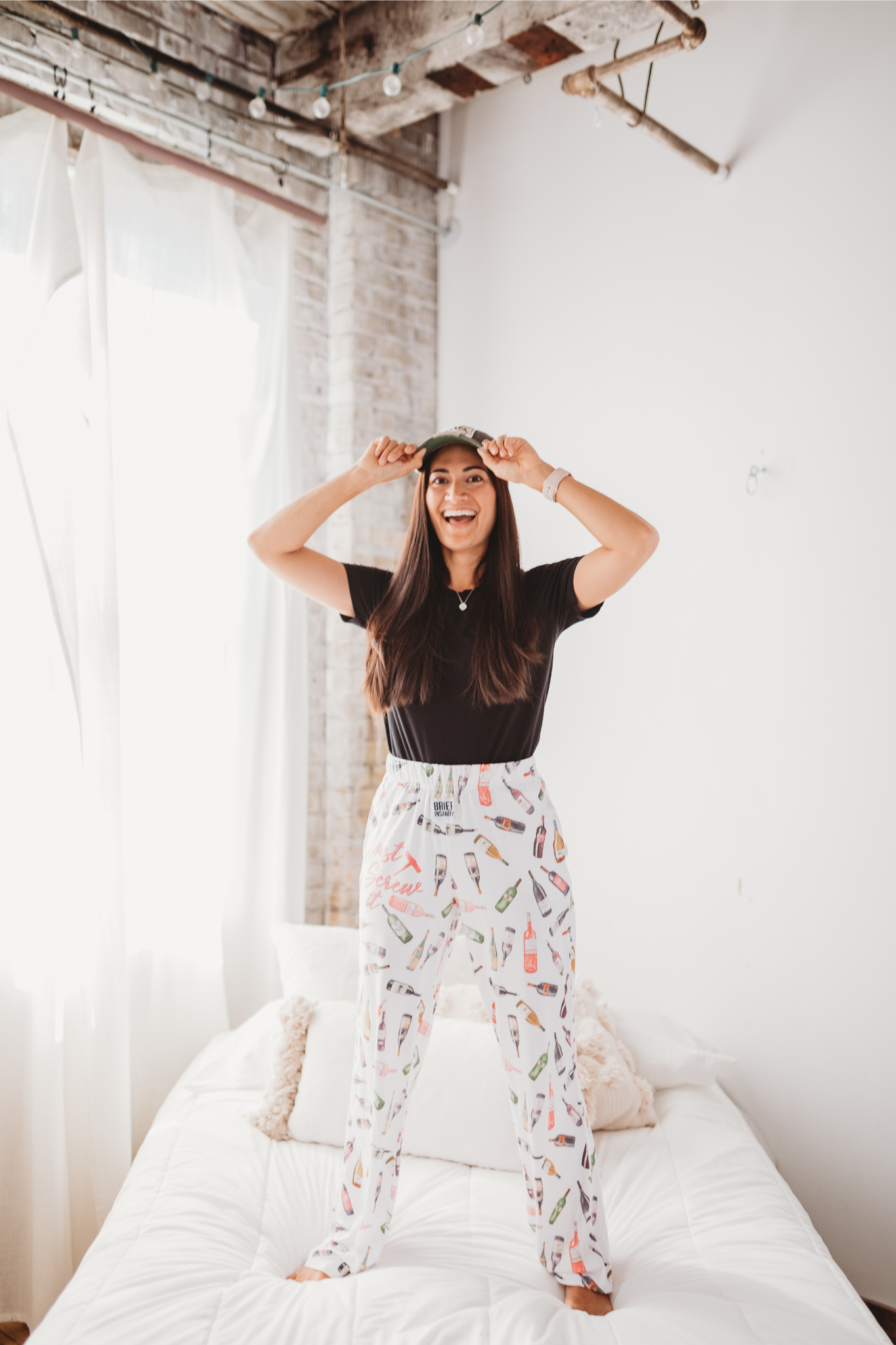 Female model standing on a white bed facing forward smiling wide and wearing Just Screw It Wine pajama lounge pants