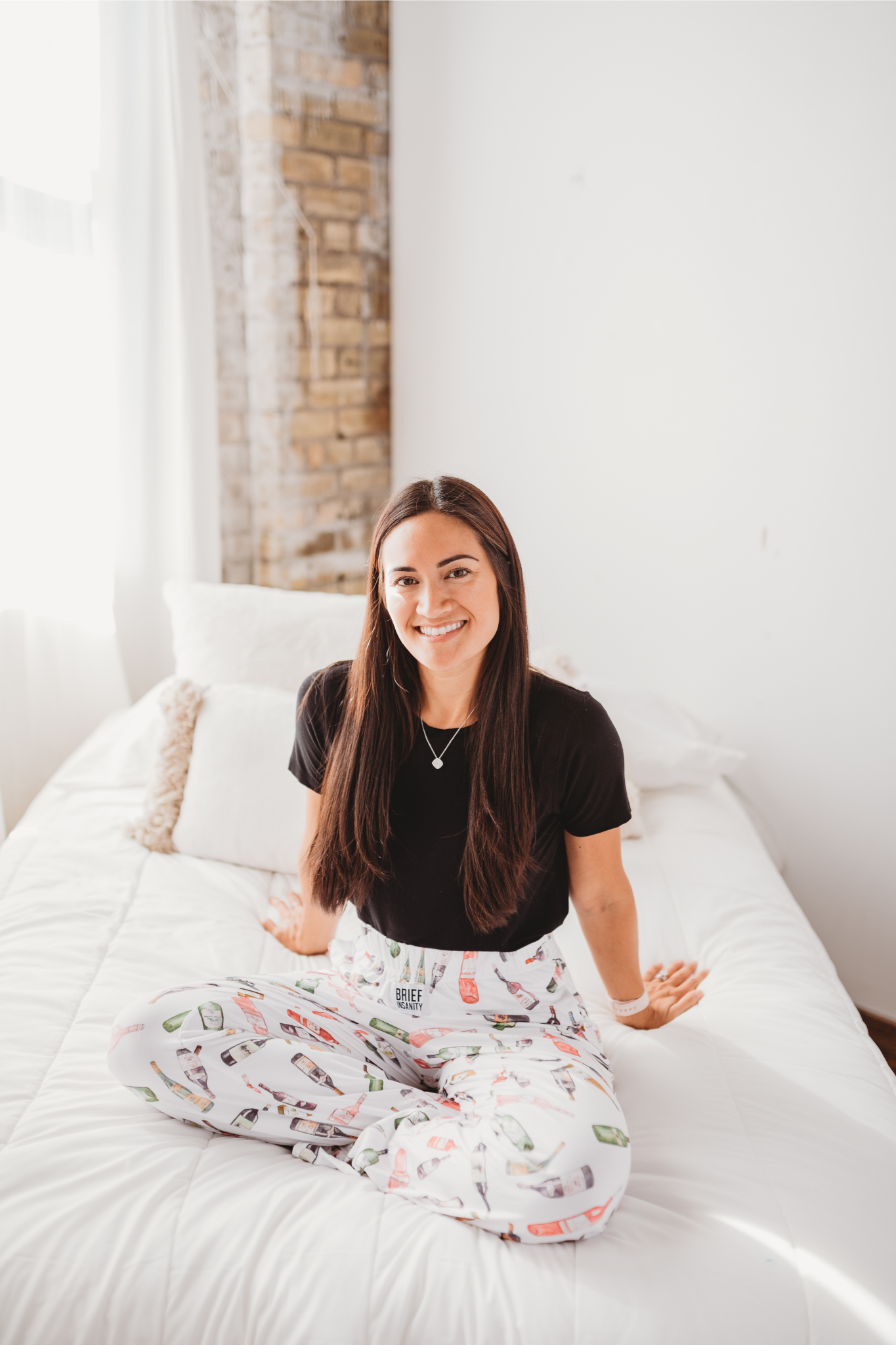 Female model sitting on white bed facing forward and smiling wearing Just Screw It Wine pajama lounge pants