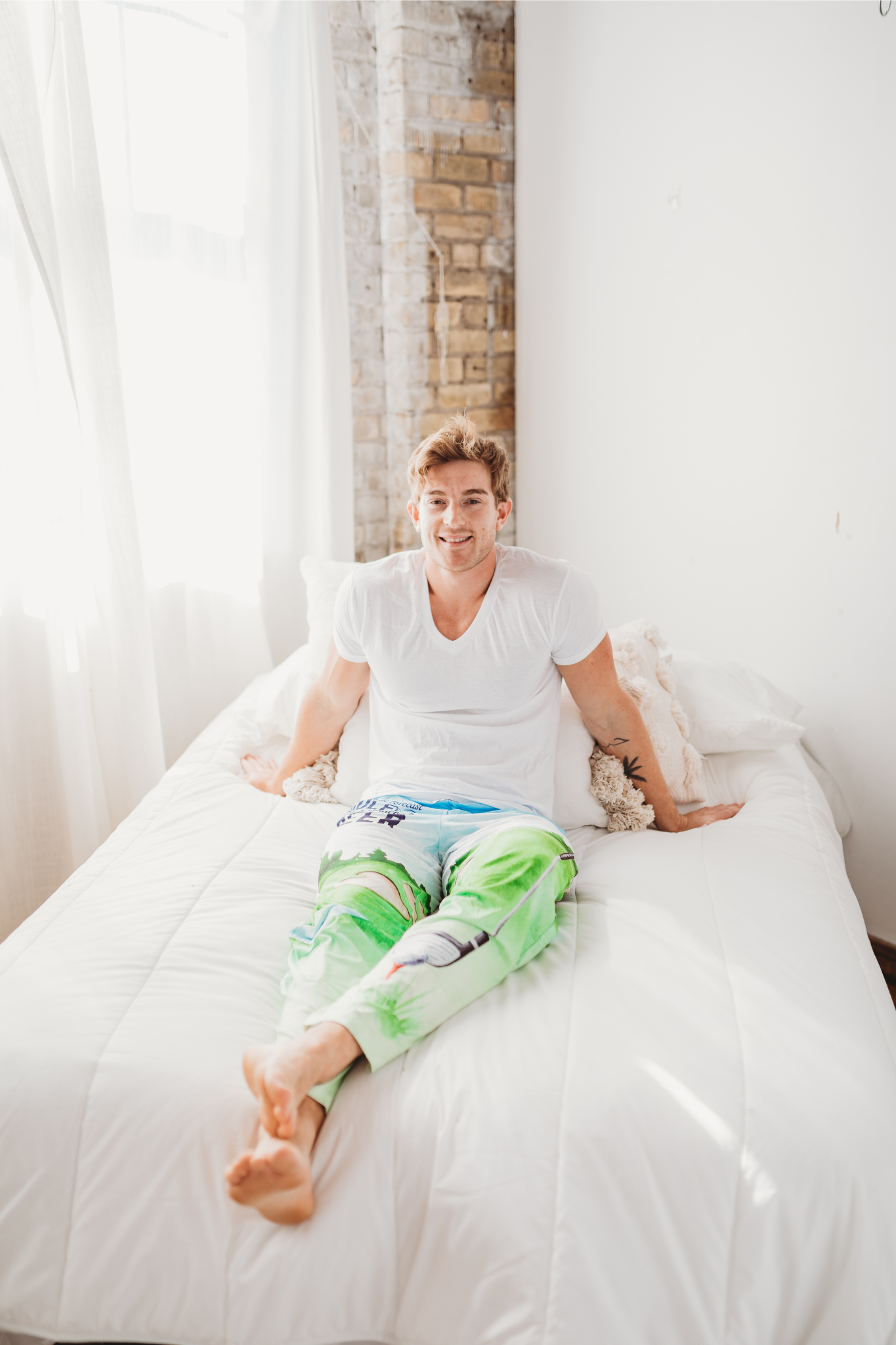 Male model sitting on a white bed facing forward with legs stretched out wearing Golf With A Chance Of Beer pajama lounge pants