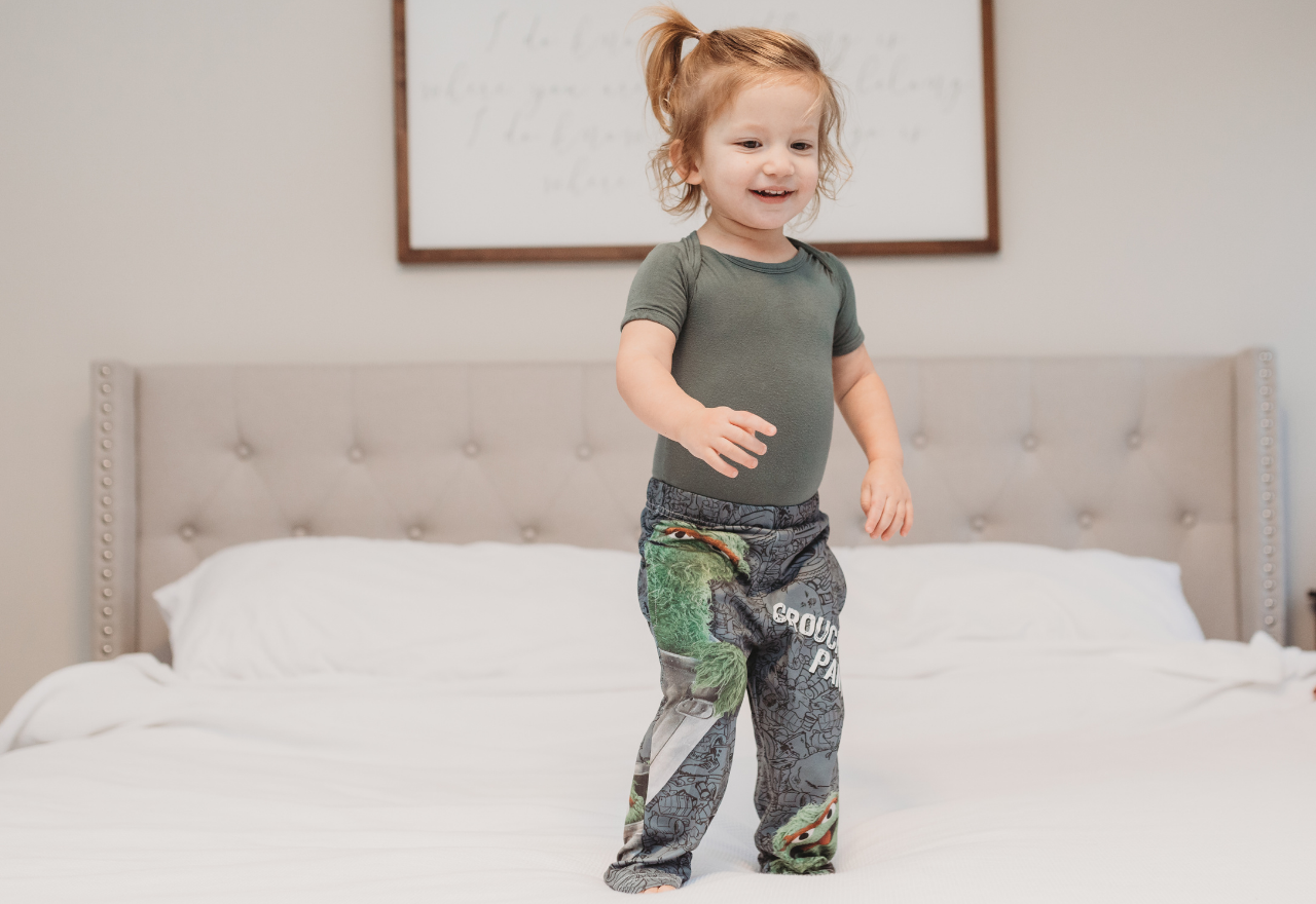 Image of little girl model standing on bed wearing Sesame Street Oscar The Grouch pajama lounge pants (front view)