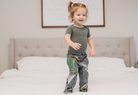 Image of little girl model standing on bed wearing Sesame Street Oscar The Grouch pajama lounge pants (front view)